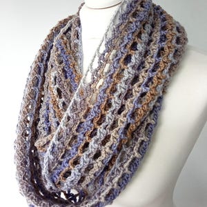 May include: A handmade infinity scarf in shades of blue, brown, and gray. The crocheted scarf is displayed on a white mannequin. The open weave design adds a delicate texture to the accessory. This versatile scarf can be worn in multiple ways.