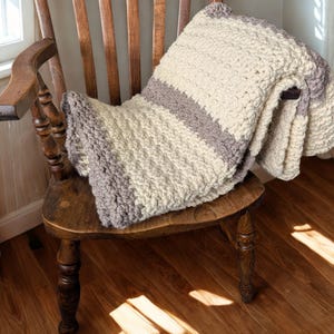 May include: A chunky knit throw blanket featuring wide horizontal stripes in cream and muted gray. The blanket is crafted with a thick, textured yarn in a consistent stitch pattern, creating a soft, folded appearance with visible color blocks.