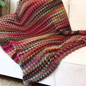 May include: A colorful, handmade crochet blanket draped over a white sofa. The blanket features a vibrant mix of colors, including shades of red, orange, brown, and teal. The textured pattern adds visual interest, making it a cozy and decorative home textile.