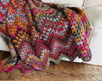 Op de afbeelding: Een handgehaakte deken met een levendig granny square patroon in een veelkleurig palet van magenta, oranje, olijfgroen, lichtblauw en donkerpaars. Het textiel heeft een getextureerd, geknoopt oppervlak met een geschulpte randafwerking.
