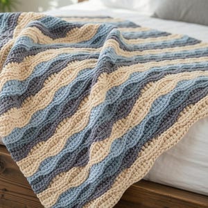 Puede incluir: Una manta tejida a ganchillo con una textura ondulada y acanalada. El diseño consiste en rayas diagonales alternas en tonos crema, azul claro y gris carbón oscuro, creando un patrón consistente en toda la superficie del tejido.