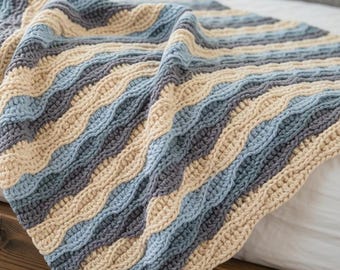 May include: A crocheted blanket featuring a wavy, rippled texture. The design consists of alternating diagonal stripes in shades of cream, light blue, and dark charcoal gray, creating a consistent pattern across the entire surface of the textile.