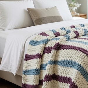 May include: A chunky knit throw blanket featuring horizontal stripes in cream, dusty blue, and deep plum. The blanket has a thick, textured yarn construction with a repeating pattern of these three colors, draped over the corner of a bed.