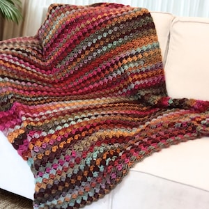 Könnte beinhalten: Eine mehrfarbige Häkeldecke, die über einem weißen Sofa drapiert ist. Die Decke zeigt eine lebendige Mischung aus Farben, darunter Rot-, Orange-, Braun- und Türkistöne. Das strukturierte Muster sorgt für visuelles Interesse und macht sie zu einem gemütlichen und dekorativen Wohnaccessoire.