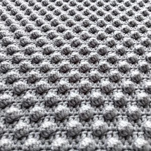 Op de afbeelding: Close-up van een grijze gehaakte deken met een textuur van bobbelsteek. Het oppervlak van de deken is dicht bedekt met gelijkmatig verdeelde, driedimensionale bobbels, waardoor een tactiel en visueel interessant ontwerp ontstaat. De kleur is een zacht, neutraal grijs.