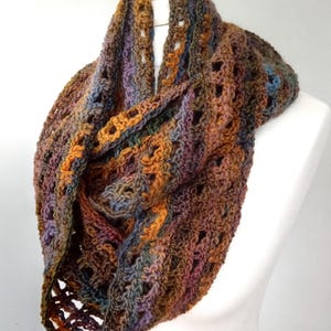 Könnte beinhalten: Handgefertigter Loop-Schal aus meliertem Garn. Der Schal hat ein Häkeldesign mit durchbrochenen Details. Die Farbpalette umfasst Brauntöne, Orange, Lila und Blau, wodurch ein warmer und strukturierter Look entsteht. Der Schal wird auf einer weißen Schaufensterpuppe präsentiert.