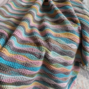 May include: A crocheted blanket featuring a repeating wavy ripple pattern. The textile is composed of horizontal stripes in various colors including soft pink, mustard yellow, light blue, sage green, grey, and cream, creating a textured surface.
