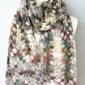 Könnte beinhalten: Handgefertigter, gehäkelter Schal im Granny-Square-Muster. Der Schal weist eine Mischung aus Farben wie Grau, Weiß, Grün und Burgunder auf. Der Schal ist über eine Schaufensterpuppe drapiert, um seine Länge und Textur zu zeigen. Ein gemütliches Accessoire.