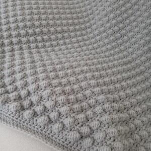 Op de afbeelding: Een close-up van een grijze, getextureerde, gehaakte deken. De deken heeft een herhalend patroon van verhoogde bobbles, waardoor een zacht, tastbaar oppervlak ontstaat. De randen zijn netjes afgewerkt met een gehaakte rand. De deken is lichtgrijs.