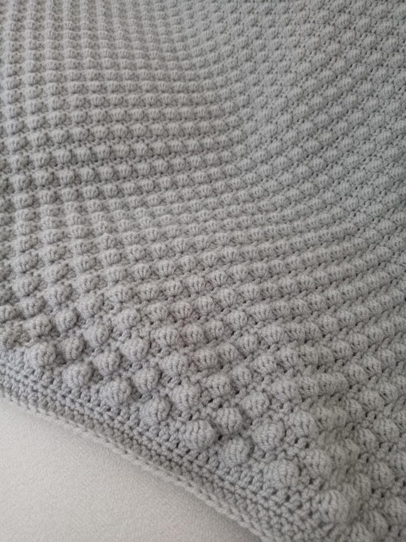 Op de afbeelding: Een close-up van een grijze, getextureerde, gehaakte deken. De deken heeft een herhalend patroon van verhoogde bobbles, waardoor een zacht, tastbaar oppervlak ontstaat. De randen zijn netjes afgewerkt met een gehaakte rand. De deken is lichtgrijs.
