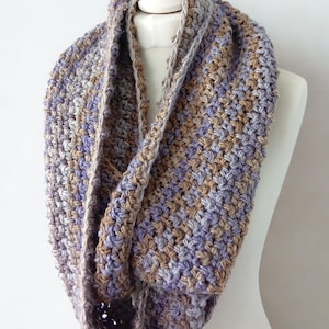 May include: A handmade infinity scarf in a soft, chunky knit. The scarf features a blend of muted colors, including lavender, tan, and gray. The scarf is displayed on a white mannequin, showcasing its circular design and textured pattern.