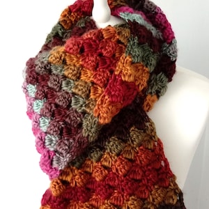 May include: A handmade crochet scarf in a variety of warm colors, including red, orange, green, and purple. The scarf is displayed on a white mannequin and features a textured, patterned design, perfect for fall or winter wear.