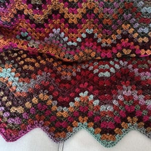 May include: A colourful, handmade crochet blanket with a scalloped edge. The blanket has a patchwork design in shades of red, pink, orange, brown, and teal. The yarn appears to be a soft, textured material.