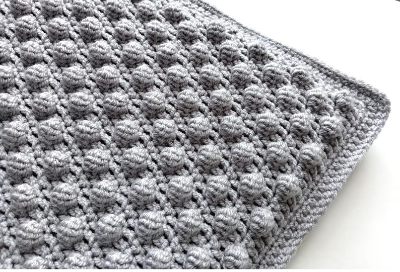 Op de afbeelding: Een close-up van een grijze gehaakte deken met een gestructureerd, verhoogd bubbelsteekpatroon. De deken is effen grijs van kleur en heeft een afgewerkte rand. De handgemaakte deken is een gezellig woondecoratie-item.