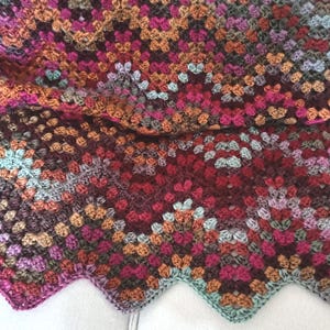 May include: A handmade crochet blanket in a granny square pattern. The blanket features a variety of colors, including shades of red, pink, orange, brown, and teal. The edges have a scalloped design.