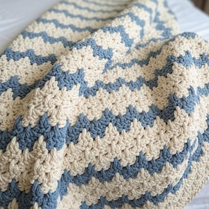 May include: A hand-crocheted blanket featuring a repeating chevron pattern in alternating stripes of dusty blue and cream-colored yarn. The textile displays a thick, textured stitch construction with a soft, folded drape across the entire surface.