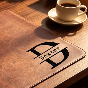 Puede incluir: Un protector de escritorio rectangular de cuero marrón con una superficie texturizada y un diseño de monograma negro con el nombre Dexter impreso en el centro. El protector está colocado sobre un escritorio de madera junto a un portátil.