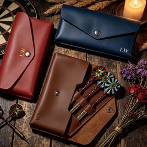 May include: Three rectangular leather dart cases in red, navy blue, and brown, each featuring a metal snap closure and the initials LW. The brown case is open, revealing three darts with colourful patterned flights and metallic barrels inside.