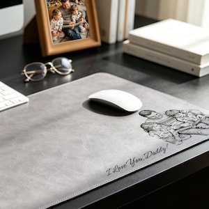 May include: A rectangular gray desk mat with a smooth, textured surface. The mat features a black line-art illustration of a parent and two children, accompanied by the printed text "I Love You, Daddy!" in a cursive script font on the lower right side.