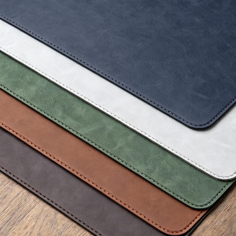 May include: A stack of five rectangular desk mats with rounded corners, featuring a textured surface. The mats are layered in colors of dark blue, white, sage green, tan, and dark brown, each finished with a matching stitched border edge.