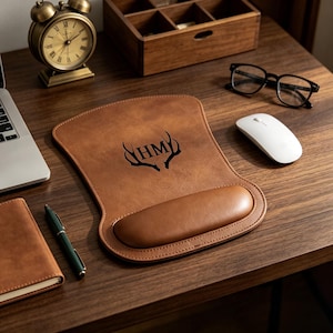 May include: A tan leather mouse pad with a built-in wrist rest, featuring a black graphic of deer antlers surrounding the letters HM. The mouse pad has a smooth, contoured shape with stitched edges, providing a functional surface for computer use.