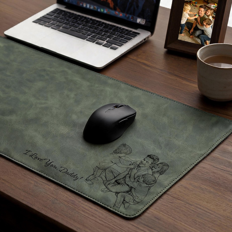 May include: A rectangular olive green desk mat featuring a laser-engraved illustration of a parent embracing two children. The text "I Love You, Daddy!" is engraved in a cursive script on the lower left side of the mat, which has a smooth texture.