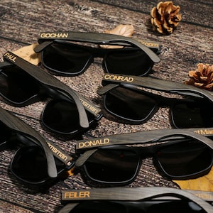 May include: Several pairs of dark wooden sunglasses with black lenses are arranged on a wooden surface. Each pair has a name engraved on the arm, including "GOCHAN", "PAUL", "CONRAD", "DONALD", and "FEILER".
