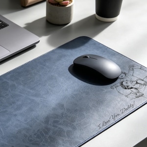 May include: A rectangular blue desk mat with a textured surface featuring a black line art illustration of a person carrying a child on their shoulders and the text "I Love You, Daddy!" printed in a cursive script font on the bottom right corner.
