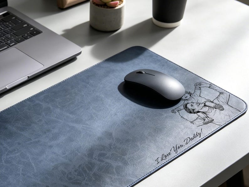 May include: A rectangular blue desk mat with a textured surface featuring a black line art illustration of a person carrying a child on their shoulders and the text "I Love You, Daddy!" printed in a cursive script font on the bottom right corner.