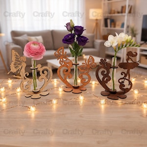 May include: Three decorative wooden stands with glass test tube vases, each featuring intricate laser-cut designs including a fairy, butterflies, and a ballerina. The stands hold a pink ranunculus, purple eustoma, and white freesia flowers.