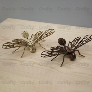 May include: Two insect sculptures crafted from laser-cut wood, one in a light natural finish and the other in a dark brown finish. Each figure features intricate, open-work wing patterns, segmented bodies, and thin legs, displaying a detailed design.