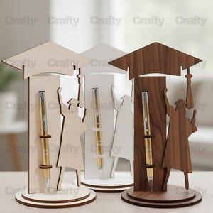 May include: Three graduation-themed pen holders made of wood in light, white, and dark brown finishes. Each holder features a flat base, a vertical panel with a gold-toned pen, and a silhouette of a graduate wearing a cap and gown.
