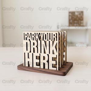 May include: Wooden drink coasters with the text "PARK YOUR DRINK HERE" cut out. The coasters are light brown and sit in a dark brown wooden holder. A second, similar item is visible in the background.