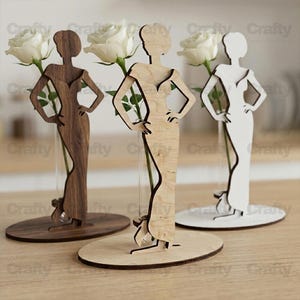 May include: Three wooden vase displays featuring a cutout silhouette of a person in a gown, each holding a single white rose in a clear glass tube. The set includes one dark brown, one light natural wood, and one white painted wooden figure.