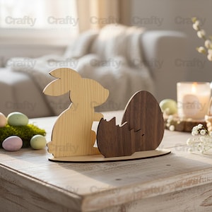 May include: Wooden Easter decoration featuring a bunny silhouette and a cracked egg. The bunny is light-colored, while the egg is a darker wood. The base is a matching wood. The word "Crafty" is printed in the background.