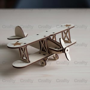 Puede incluir: Un modelo de avión biplano de madera con hélice, ruedas y timón de cola. El modelo es de color marrón claro con detalles en marrón oscuro. Las alas presentan recortes de estrellas. La palabra "Crafty" está impresa en la superficie.