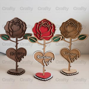 Könnte beinhalten: Drei lasergeschnittene Rosenornamente aus Holz auf Ständern. Jedes zeigt eine Rosenblüte, grüne Blätter und ein herzförmiges Schild mit der Gravur Katrina love. Die mittlere Rose ist rot, die anderen sind in Naturholztönen mit dunklen Akzenten.