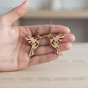May include: A pair of wooden hummingbird earrings. The earrings are laser-cut with intricate details, showcasing the bird's wings and body. The earrings hang from silver-colored ear wires, and are held in a hand.