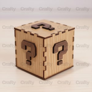 May include: A wooden cube-shaped box with a question mark on each side. The box is made of light-colored wood with darker brown question marks. The box has a laser-cut design and is likely a decorative or storage item.
