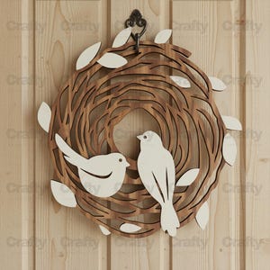 May include: A circular wall hanging featuring a brown, laser-cut wooden nest design with two white birds and several white leaves attached. The piece is suspended from a dark metal hook at the top, showcasing a layered, cutout aesthetic.