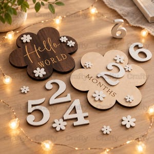 May include: Two flower-shaped wooden plaques with white floral accents. One dark brown plaque features the text Hello World. One light wood plaque features the text MONTHS. Includes separate white wooden numbers 2, 4, 5, and 6 for milestone tracking.