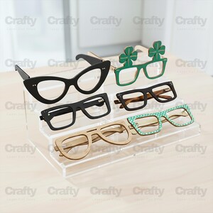 May include: A clear acrylic tiered display stand holding five pairs of glasses. The collection includes black cat-eye frames, green shamrock-shaped frames, black rectangular frames, a natural wood-toned pair, and a pair with a green polka-dot pattern.