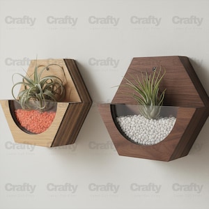 Może przedstawiać: Dwie sześciokątne drewniane donice ścienne z przezroczystymi panelami przednimi. Jedna donica ma pomarańczowe kamyki i roślinę, druga białe kamyki i roślinę. Donice są zamontowane na białej ścianie.