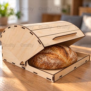 May include: A light-coloured wooden bread box with a curved, slatted roll-top lid and a flat base. A round, crusty loaf of bread sits inside the open container. The wood features visible grain patterns and dark, laser-cut edges throughout.