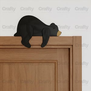 May include: A black wooden bear decoration, designed to rest on a door frame. The bear is in a sleeping position, with a light brown nose. The door frame is made of wood.