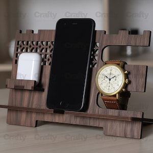 Peut inclure: Un organiseur de bureau en bois brun foncé. Il comporte des emplacements pour un smartphone, des écouteurs blancs et une montre-bracelet avec un boîtier doré et un bracelet en cuir marron. L'organisateur a un motif découpé décoratif.