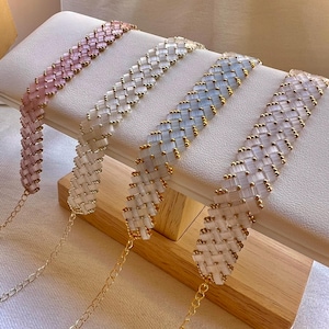 Op de afbeelding: Vier kralen chokers in pastelkleuren: roze, wit, lichtblauw en lavendel. Elke choker heeft een geweven patroon van rechthoekige kralen en kleine goudkleurige kralen. De chokers hebben delicate goudkleurige kettingverlengingen met een kleine bedel.