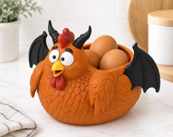 Könnte beinhalten: Eine orangefarbene Schale in Hühnerform mit schwarzen Fledermausflügeln und Hörnern, einem roten Kamm und Kehllappen, einem gelben Schnabel und großen weißen Augen. Die Schale enthält drei braune Eier, der Körper ist federartig strukturiert.