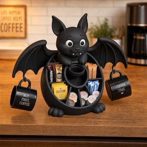 May include: A black bat-shaped coffee station organizer with a central compartment for tea bags, sugar sticks, and creamer pods. Two black mugs hang from the bat's wings. The left mug reads "BUT FIRST, COFFEE" and the right mug reads "COFFEE TIME".