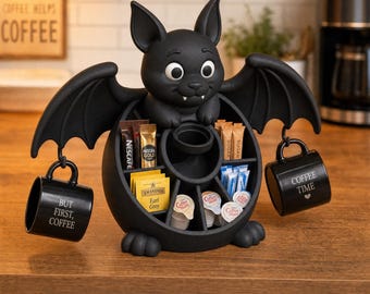 Könnte beinhalten: Ein schwarzer Kaffee-Organizer in Fledermausform mit einem zentralen Fach für Teebeutel, Zuckersticks und Kaffeeweißer-Portionen. Zwei schwarze Tassen hängen an den Flügeln. Die linke Tasse trägt die Aufschrift "BUT FIRST, COFFEE".